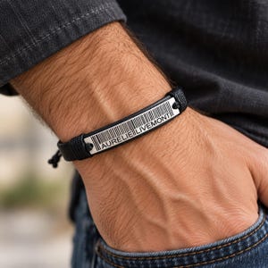 Puede incluir: Una pulsera de cordón negro con una placa rectangular de metal plateado. La placa está grabada con un gráfico de código de barras y el texto AURELIE LIVEMONT en letras mayúsculas negras. La pulsera tiene extremos de cordón ajustables.