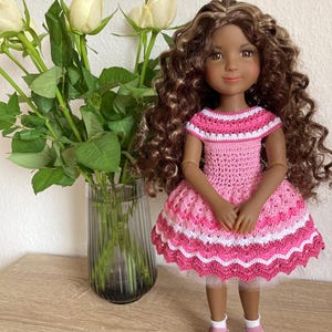 Op de afbeelding: Pop met bruin krullend haar in een roze en witte gehaakte jurk en roze schoenen. De pop staat op een houten oppervlak naast een vaas met witte rozen.
