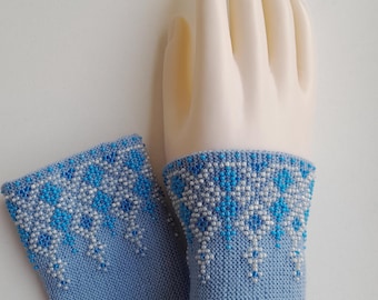 May include: Light blue knitted wrist warmers with a beaded pattern. The top edge has a design of white and blue beads, with a cascading effect. The wrist warmers are displayed on a mannequin hand.