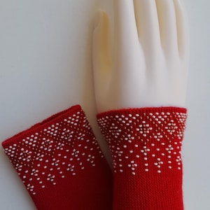 May include: A pair of red knitted wrist warmers with a white and brown beaded pattern. One wrist warmer is on a mannequin hand, the other is next to it. The wrist warmers are a cozy accessory.