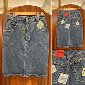Peut inclure: Jupe crayon en jean bleu moyen avec plusieurs écussons et étiquettes. La jupe a une conception classique à cinq poches. Les écussons présentent du texte et des graphiques. La jupe est présentée sur un cintre en bois.