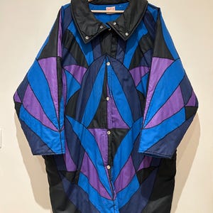 May include: A vintage long coat with a geometric pattern in blue, purple, and black. The coat has a black collar and is buttoned up. The coat is hanging on a wooden hanger.