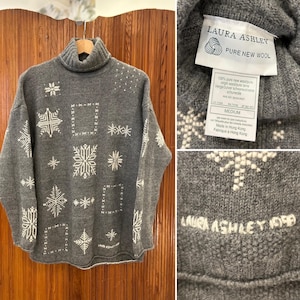 Può includere: Maglione grigio a collo alto con un motivo a fiocco di neve e il testo "LAURA ASHLEY PURE NEW WOOL" sull'etichetta. L'etichetta riporta anche "MEDIUM" e "Made in Hong Kong".