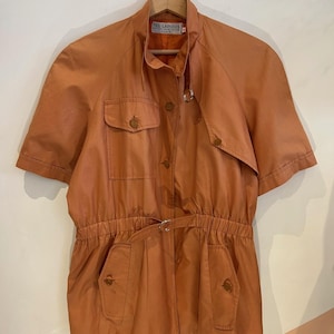 May include: A short-sleeved, burnt orange jacket featuring a high collar, a button-front closure, and a cinched waist with a belt. The garment includes two chest pockets and two lower pockets, all with button flaps. A label reads TED LAPIDUS.