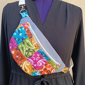 Peut inclure: Un sac banane coloré avec un motif patchwork. Le corps principal présente un mélange vibrant de carrés à motifs dans les tons orange, rose, bleu et vert. Il a une base grise semblable à du denim et une fermeture éclair argentée. La sangle réglable est blanche.