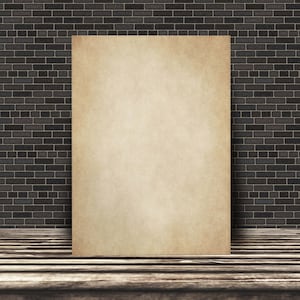 May include: A blank, aged, cream-colored paper poster is leaning against a dark brick wall. The poster is in front of a wooden floor.