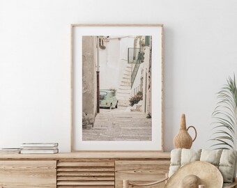 May include: A framed print of a European street scene. The image features a light green vintage car parked on a cobblestone street. The scene is framed by white buildings and a staircase. The print is displayed on a wooden cabinet.
