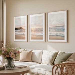 May include: Three framed beach landscape prints with a neutral color palette hang above a white sofa. The prints depict ocean waves, sand, and a sunset. The room also features a round wooden coffee table, a vase of pink flowers, and woven baskets.