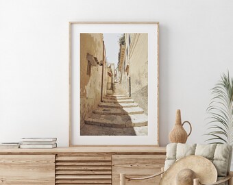 May include: A framed photograph of a narrow, sunlit street in a European city. The street is lined with buildings and has a set of stone steps leading up to the top of the frame.