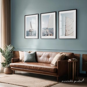 May include: Three framed prints of sailboats at sea. The prints are in black frames and hang on a blue wall above a brown leather sofa. The images depict sailboats with white sails on a blue ocean.