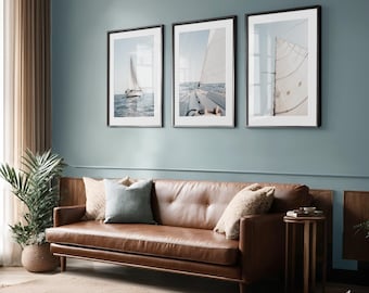 May include: Three framed prints of sailboats at sea. The prints are in black frames and hang on a blue wall above a brown leather sofa. The images depict sailboats with white sails on a blue ocean.