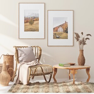 May include: Two framed landscape photographs of a mountain scene with a single tree in the foreground and a house in a valley with trees in the background. The photographs are hanging on a light beige wall above a wicker chair with a light beige throw blanket and a light beige pillow. A wicker table with a green book and a wooden bowl is in front of the chair. A wicker vase with dried flowers is on the right side of the image.