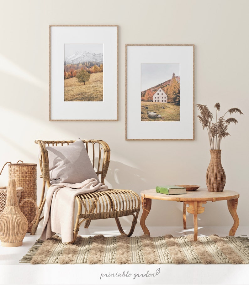May include: Two framed landscape photographs of a mountain scene with a single tree in the foreground and a house in a valley with trees in the background. The photographs are hanging on a light beige wall above a wicker chair with a light beige throw blanket and a light beige pillow. A wicker table with a green book and a wooden bowl is in front of the chair. A wicker vase with dried flowers is on the right side of the image.