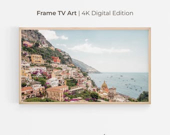 May include: A framed digital print of a coastal Italian town. The image features colorful buildings cascading down a hillside towards the sea. The sky is a pale blue, and the frame is a light wood color. Text at the top reads "Frame TV Art | 4K Digital Edition".