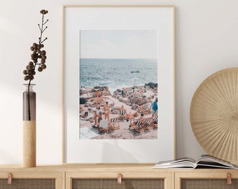 May include: A vintage photograph of a beach scene with people relaxing on striped beach chairs. The photo is framed in a light wood frame and is hanging on a white wall.