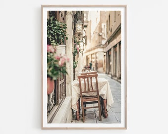 May include: A vintage-style photograph of a cafe table and chairs set up outside on a cobblestone street in a European city. The table is covered with a white tablecloth and the chairs are wooden. The street is lined with buildings and there are flowers in pots hanging from the buildings.