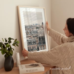 Peut inclure: Une impression encadrée d'un paysage urbain avec des bâtiments aux toits gris et un ciel nuageux. L'impression est tenue par une personne portant un pull beige. Un vase avec de la verdure et un livre sont sur une surface en bois.