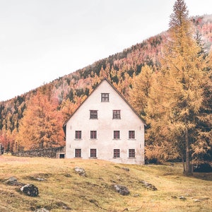 May include: A white house with multiple windows sits in a clearing surrounded by trees with fall foliage. The house has a triangular roof and a stone foundation.