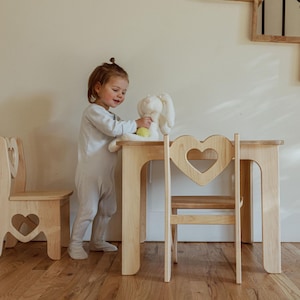 May include: A light-colored wooden children's table and chair set. The chair backs and table have heart-shaped cutouts. A child is standing next to the table with a stuffed bunny. The set is made of light-colored wood.