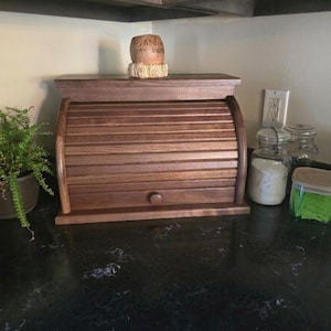 May include: A dark brown wooden bread box with a roll-top tambour door and a small circular knob. The bread box features horizontal slats and a flat top surface. A small, light-colored decorative object with handwritten text sits on top.
