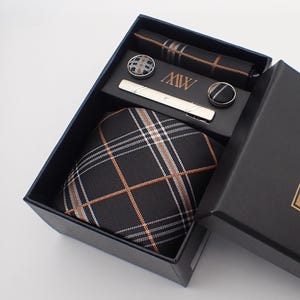 May include: A black gift box containing a tie, pocket square, cufflinks, and tie clip. The tie and pocket square have a black, white, and orange plaid design. The box has a gold label with the initials "MW".