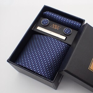 Op de afbeelding: Een zwarte geschenkdoos met een blauwe en witte gepatenteerde stropdas, een stropdasclip en manchetknopen. De doos heeft de tekst "MW by BOWTIE SUPPLY" op de voorkant.