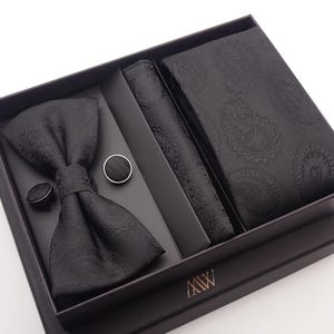 May include: A black formal accessory set presented in a gift box. The set includes a bow tie, cufflinks, a pocket square, and a necktie, all featuring a textured paisley design. The box is black with the initials "MW".
