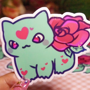 Op de afbeelding: Een sticker met een stripfiguur met een mintgroen lichaam, roze hartjes en een grote roze roos. Het personage heeft roze ogen en blos. De sticker wordt in een hand gehouden, met andere soortgelijke stickers op de achtergrond.