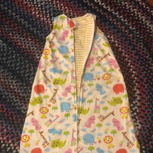 May include: White sleeping bag with a zipper down the front. The sleeping bag has a colorful pattern of animals, including giraffes, elephants, lions, and dinosaurs. The sleeping bag is lined with yellow and white striped fabric.