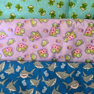 Puede incluir: Se muestran tres patrones de tela. La tela superior es azul claro con ilustraciones de ranas verdes. La tela del medio es lavanda con ilustraciones de tortugas. La tela inferior es azul con ilustraciones de tiburones.