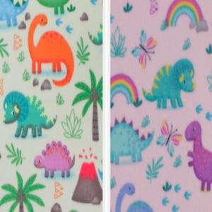 Puede incluir: Dos muestras de tela con patrones de dinosaurios. La primera muestra es verde con fondo blanco y presenta dinosaurios, palmeras y volcanes. La segunda muestra es rosa con fondo blanco y presenta dinosaurios, arcoíris y mariposas.