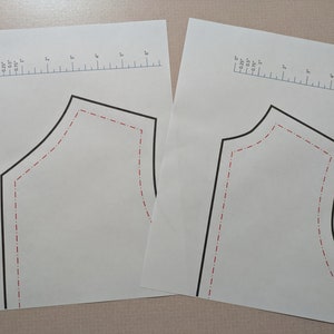 May include: Two white paper pattern pieces with black outlines and red dashed inner lines. Each has a ruler printed along the top edge, marked in inches. The patterns are for sewing or crafting projects.