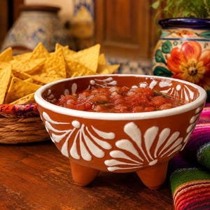 May include: A terracotta bowl with three short legs and white hand-painted floral patterns filled with red salsa. The bowl is positioned near a woven basket filled with triangular yellow corn tortilla chips and a colorful ceramic jar with floral designs.