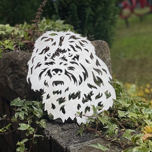 May include: White metal cutout of a dog's head with curly fur. The dog is looking directly at the viewer with its mouth closed. The cutout is placed on a rock with green foliage in the background.