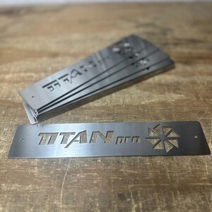 Może przedstawiać: Stos prostokątnych metalowych płytek z wyciętym napisem "TITAN pro". Srebrne płytki mają geometryczny wz&oacute;r i leżą na drewnianej powierzchni.