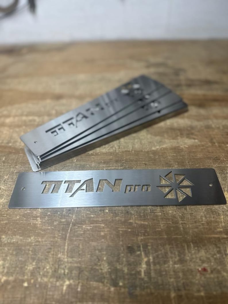 Może przedstawiać: Stos prostokątnych metalowych płytek z wyciętym napisem "TITAN pro". Srebrne płytki mają geometryczny wz&oacute;r i leżą na drewnianej powierzchni.