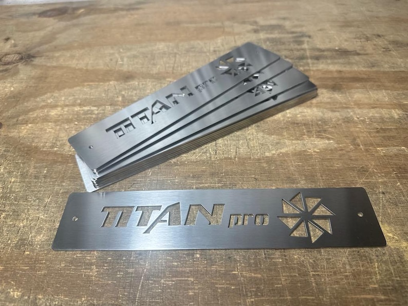 Może przedstawiać: Prostokątne płyty ze szczotkowanego metalu z wyciętym napisem "TITAN pro". Widoczny stos płyt. Płyty mają geometryczny wz&oacute;r po prawej stronie. Na drewnianej powierzchni.