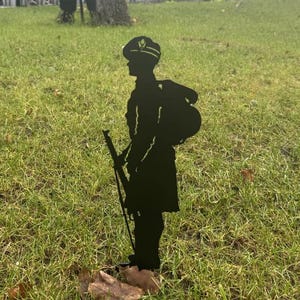 May include: Black metal silhouette of a soldier standing to attention, holding a rifle. The soldier is wearing a hat with a gold badge. The silhouette is placed on a grassy lawn.