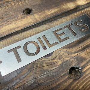 Może przedstawiać: Prostokątny, szczotkowany metalowy znak z wyciętym słowem "TOILETS" wielkimi literami. Znak ma otw&oacute;r w lewym g&oacute;rnym rogu do zawieszenia. Znak jest umieszczony na rustykalnym, drewnianym tle.