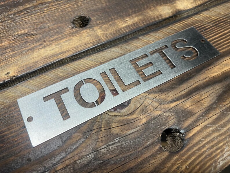 Może przedstawiać: Prostokątny, szczotkowany metalowy znak z wyciętym słowem "TOILETS" wielkimi literami. Znak ma otw&oacute;r w lewym g&oacute;rnym rogu do zawieszenia. Znak jest umieszczony na rustykalnym, drewnianym tle.