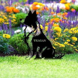 May include: A black metal dog silhouette with the name "TEAGAN" in white, set in a garden. The dog is in front of a colourful flower bed with yellow, orange, purple, and white flowers, against a backdrop of green grass.