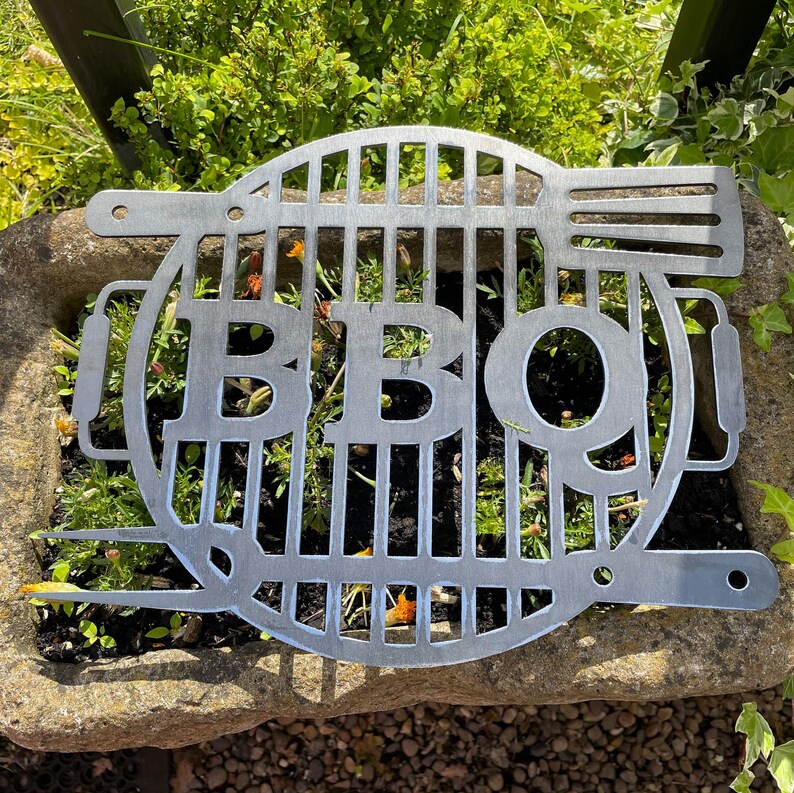 May include: A metal BBQ grill with the word "BBQ" cut out in the centre. The grill is sitting on a stone surface.