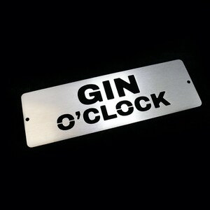 May include: A silver metal sign with the text "GIN O'CLOCK" in black lettering.