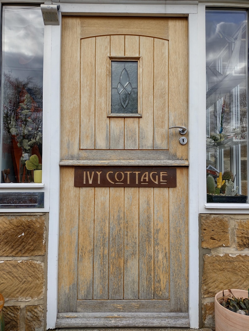 Może przedstawiać: Zniszczone drewniane drzwi z małym witrażowym oknem. Metalowy znak nad drzwiami głosi "IVY COTTAGE". Okna z białymi ramami i kamienne ściany flankują drzwi.
