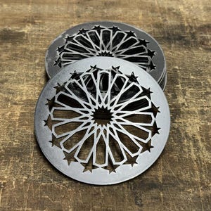 Könnte beinhalten: Ein Stapel silberfarbener Metalluntersetzer mit einem geometrischen Sternmuster. Die Untersetzer haben eine runde Form mit aufwendigen Ausschnitten. Der Hintergrund ist eine rustikale Holzoberfläche.