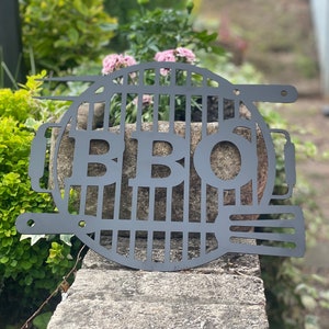 May include: Black metal BBQ grill sign with the letters "BBQ" cut out of the grill design. The sign is hanging on a stone wall with green plants in the background.