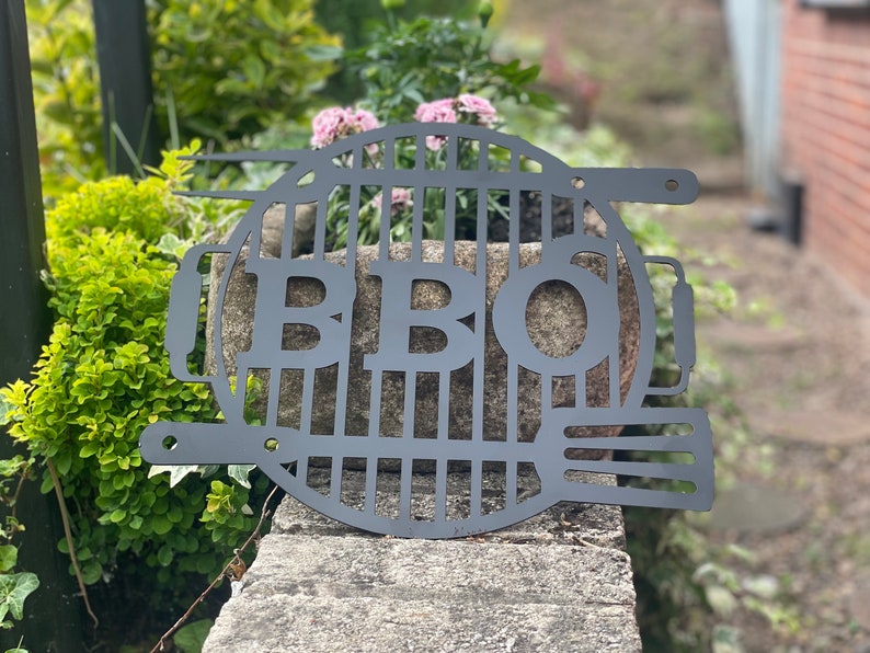 May include: Black metal BBQ grill sign with the letters "BBQ" cut out of the grill design. The sign is hanging on a stone wall with green plants in the background.