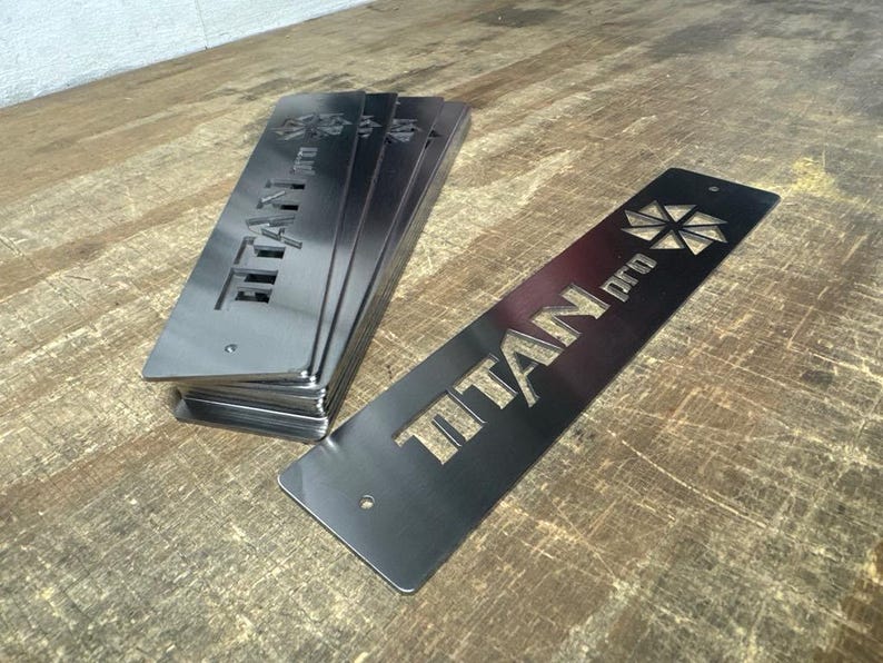 Może przedstawiać: Stos prostokątnych, metalicznych, szarych metalowych płyt z wyciętym tekstem "TITAN pro" i geometrycznym wzorem. Płyty leżą na drewnianej powierzchni.