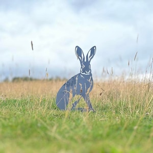 May include: A metal silhouette of a hare, standing in a field of long grass. The hare is facing forward with its ears perked up.