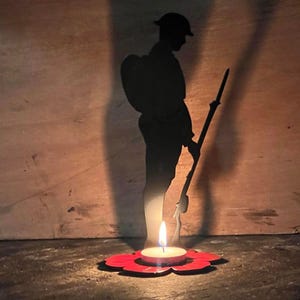 May include: A silhouette of a soldier holding a rifle stands in front of a lit candle in a red poppy-shaped holder. The soldier's shadow is cast on a wooden surface.